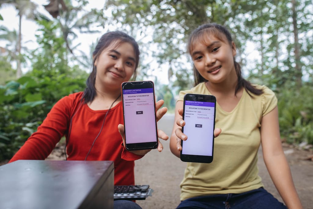 A girl in a red top and a girl in a green top hold up two cellphones.
